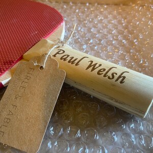 May include: A red ping pong paddle with a wooden handle engraved with the name "Paul Welsh." A brown tag is attached with the words "Firle & Fable" printed on it. The paddle is resting on bubble wrap.