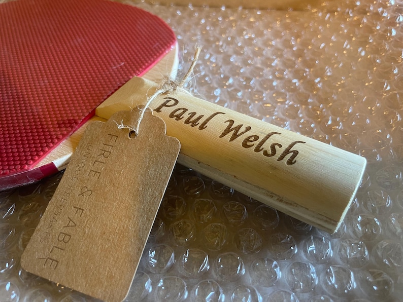 May include: A red ping pong paddle with a wooden handle engraved with the name "Paul Welsh." A brown tag is attached with the words "Firle & Fable" printed on it. The paddle is resting on bubble wrap.
