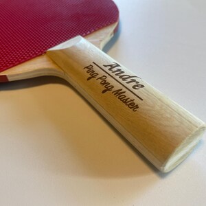 May include: A ping pong paddle with a red rubber surface and a light wood handle. The handle is engraved with the name "Andre" and the words "Ping Pong Master".