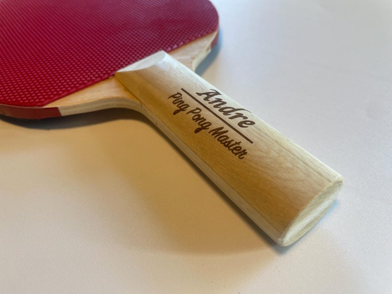 May include: A ping pong paddle with a red rubber surface and a light wood handle. The handle is engraved with the name "Andre" and the words "Ping Pong Master".