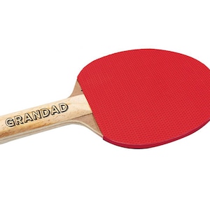 Puede incluir: Una raqueta de ping pong con superficie de goma roja y mango de madera. El mango es de madera clara con la palabra "GRANDAD" impresa en letras negras. Dise&ntilde;ada para jugar al tenis de mesa.