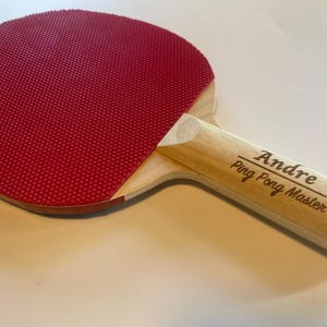 May include: A ping pong paddle with a red rubber surface and a light wood handle. The handle is engraved with the name "Andre" and the text "Ping Pong Master". The paddle is designed for table tennis.