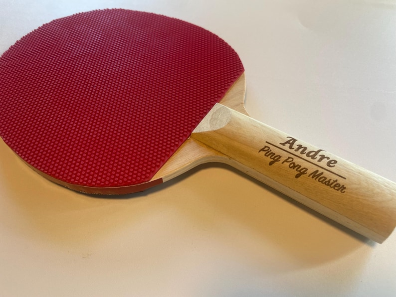 May include: A ping pong paddle with a red rubber surface and a light wood handle. The handle is engraved with the name "Andre" and the text "Ping Pong Master". The paddle is designed for table tennis.