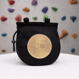 May include: Black chalk bag with a round, tan patch featuring a climber graphic and the initials "JB." The bag has a drawstring closure and two loops. The background shows a climbing wall with colourful holds.