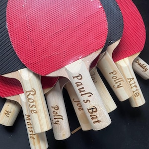 Puede incluir: Varias palas de ping-pong con mangos de madera y superficies rojas y negras. Los mangos est&aacute;n grabados con nombres como "Paul's Bat", "Polly" y "Alfie".