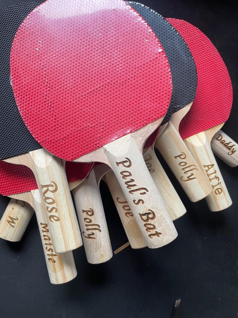Puede incluir: Varias palas de ping-pong con mangos de madera y superficies rojas y negras. Los mangos est&aacute;n grabados con nombres como "Paul's Bat", "Polly" y "Alfie".