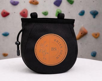 May include: Black chalk bag with a drawstring closure and two loops. A circular brown leather patch features a climber graphic and the text "BRIAN SHAW" and "BS". The bag is designed for rock climbing.
