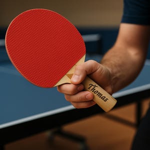 Puede incluir: Una raqueta de ping pong roja con mango de madera. La raqueta tiene una superficie de goma roja texturizada y un mango de madera de color claro. El mango est&aacute; grabado con el nombre "Thomas".