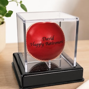 Puede incluir: Una pelota de críquet roja con la inscripción "David Happy Retirement" se exhibe en una caja de acrílico transparente sobre una base negra. Se aprecia la costura de la pelota. Un regalo de jubilación único.