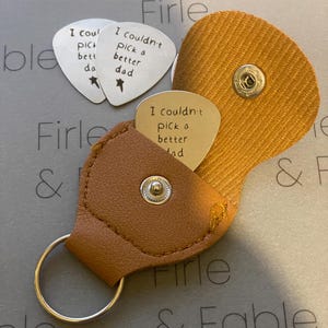May include: A brown leather guitar pick holder with a key ring. The holder has a flap that opens to reveal three silver guitar picks with the inscription "I couldn't pick a better dad" on two of the picks.