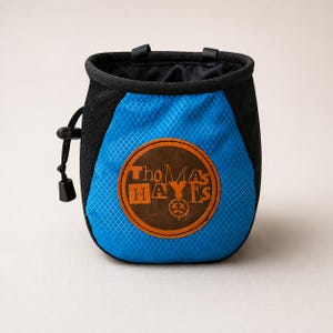 Może przedstawiać: Niebiesko-czarny worek na magnezję wspinaczkową z rombowym wzorem. Worek posiada zamknięcie na sznurek i brązową skórzaną naszywkę z pomarańczowym napisem "Thomas Hayes". Dwa czarne pętle u góry.