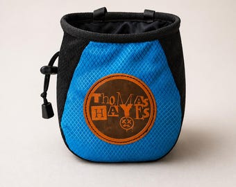 May include: A blue and black climbing chalk bag with a diamond pattern. The bag has a drawstring closure and a brown leather patch with the text "Thomas Hayes" in orange. Two black loops are at the top.