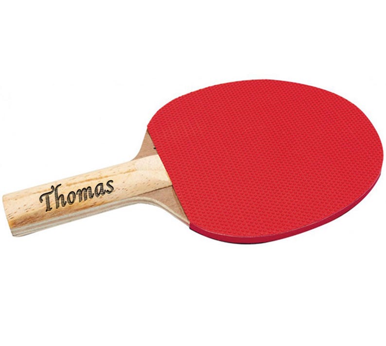 Puede incluir: Una raqueta de ping pong con una superficie de goma roja y un mango de madera clara. El nombre "Thomas" est&aacute; impreso en el mango en cursiva. La raqueta est&aacute; dise&ntilde;ada para jugar al tenis de mesa.