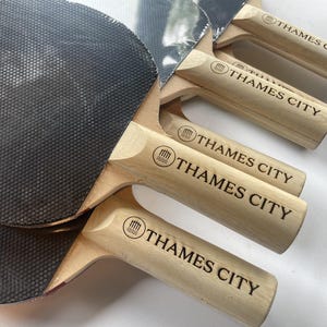 Könnte beinhalten: Ein Set aus vier Tischtennisschlägern mit schwarzer, strukturierter Gummioberfläche und hellen Holzgriffen. Jeder Griff ist mit einem kreisförmigen Logo versehen, das ein Brückensymbol und den Text THAMES CITY in schwarzen Großbuchstaben zeigt.
