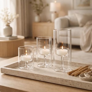 May include: Three clear glass candle holders of varying heights, each with a lit white candle. The candle holders are on a rectangular stone tray, with cinnamon sticks and small ceramic bowls. The scene is set in a living room.