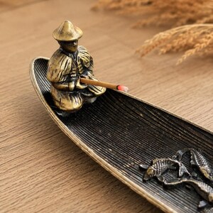 May include: A metallic incense burner shaped like a long, curved boat with a textured black interior. A bronze-toned figure wearing a conical hat sits at one end, holding a lit incense stick. Two bronze fish figures are attached to the base.