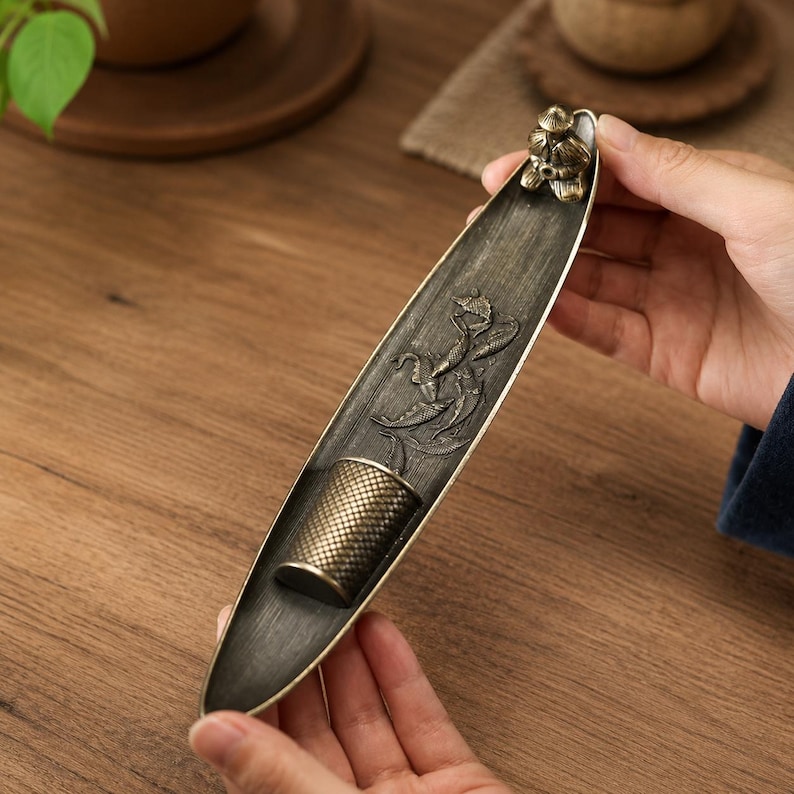 May include: A boat-shaped incense burner made of dark, brushed metal. The interior features a relief carving of fish swimming among reeds, a cylindrical holder for incense sticks, and a small figurine of a person wearing a conical hat.