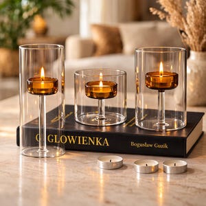 May include: Three glass candle holders with lit candles inside. The candles are in amber-colored holders, and the holders are on a black book. Three tealight candles are in front of the candle holders.