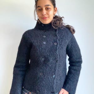 May include: A black, double-breasted jacket with a high collar and large buttons. The jacket has a textured appearance and is paired with blue jeans. Sunglasses rest on top of the jacket. The jacket is a stylish outerwear option.