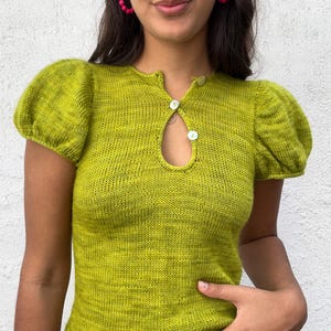 Könnte beinhalten: Ein chartreusegrünes Strickoberteil mit kurzen, gepufften Ärmeln. Das Oberteil hat einen Schlüssellochausschnitt, der mit zwei weißen Knöpfen befestigt ist. Der Strickstoff hat ein strukturiertes Aussehen.