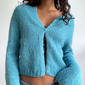 May include: A light blue, fuzzy cardigan with a V-neck and button closure. The long-sleeved sweater is made of a soft, textured material. The cardigan is paired with blue jeans.