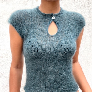 Puede incluir: Una blusa de punto de color verde azulado, de manga corta, con un detalle de ojo de cerradura y un cierre de botón único. La blusa tiene un cuello redondo y una textura ligeramente difusa. La prenda es de estilo ajustado.