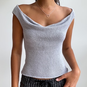 Może przedstawiać: Jasnoszary, dzianinowy top z odkrytymi ramionami i dekoltem w kształcie łódki. Top ma prążkowaną fakturę i dopasowaną sylwetkę. Widoczny jest srebrny naszyjnik z małą, okrągłą zawieszką. Osoba ma na sobie czarno-białe spodnie w paski.