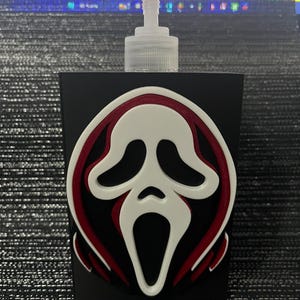 May include: A black rectangular soap dispenser featuring a prominent, raised three-dimensional Ghostface mask emblem. The mask is white with deep red accents around the eyes and mouth, centred on the front of the matte black container with a clear pump.