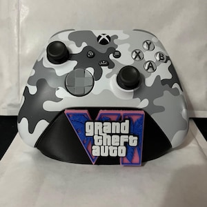May include: A white, grey, and black camouflage patterned video game controller featuring a black base stand with a pink and blue Grand Theft Auto VI logo emblem attached to the front, including standard buttons, joysticks, and a D-pad.