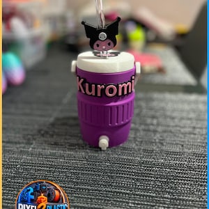 May include: A purple plastic beverage cooler with a white lid and a small white spigot at the base. The name Kuromi is displayed in pink letters on the front. A clear straw extends from the top, featuring a black Kuromi character topper.