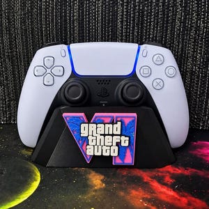 May include: A white and black wireless game controller resting on a black stand. The stand features a pink and blue graphic logo with the text Grand Theft Auto VI, including stylized palm tree silhouettes behind the Roman numeral VI.
