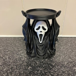 May include: A black and white candle holder featuring a figure in a hooded robe with a white ghost-like mask. The figure has its arms raised to support a circular black platform on top. The design includes detailed folds in the fabric of the robe.