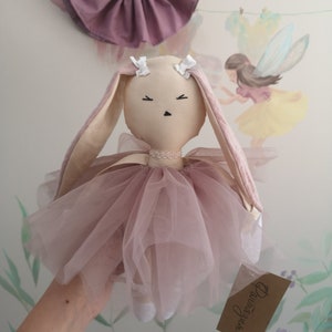 May include: A handmade pink and beige bunny doll wearing a pink tulle tutu and a white pearl necklace. The doll has a sweet expression and is holding a small brown tag that says "Dreamweaver".