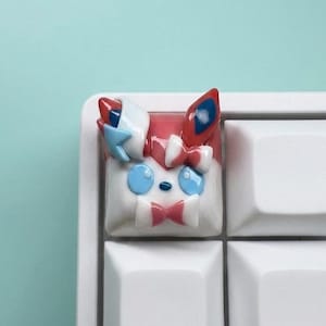 May include: A custom artisan mechanical keyboard keycap featuring a sculpted character design with a white base, light blue eyes, and pink and red accents. The keycap is shaped like a stylized creature head with ears, bows, and ribbons.