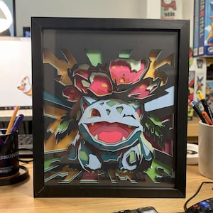 May include: A multi-layered paper shadow box art piece in a black frame featuring a stylised depiction of the creature Venusaur. The design uses depth and cutouts to create a 3D effect with green, pink, blue, and yellow colours in a comic style.