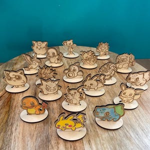 Könnte beinhalten: Holzfiguren verschiedener Pokémon-Charaktere, darunter Pikachu und Bisasam, werden auf kleinen, runden Holzsockeln präsentiert. Die Figuren sind lasergeschnitten mit detaillierten Konturen. Sie stehen auf einer Holzoberfläche vor einem türkisfarbenen Hintergrund.