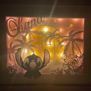 Può includere: Un'opera d'arte murale illuminata con una silhouette di Stitch di Lilo & Stitch, palme e un tramonto. La parola "Ohana" è scritta in un font decorativo sopra la scena.