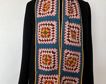 May include: A handmade crochet scarf featuring a granny square design. The scarf has a teal blue background with squares in shades of pink, burgundy, yellow, and green. The edges are trimmed with a yellow border. The scarf is displayed on a black coat.