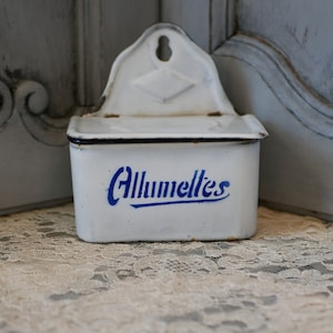May include: White enamel matchbox holder with a blue trim and the word "Allumelles" written in blue lettering.