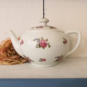 May include: White porcelain teapot with a lid, decorated with pink roses and green leaves. The teapot has a curved spout and a handle. The body of the teapot has vertical ribbed detailing. A decorative flower is next to the teapot.