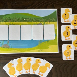Könnte beinhalten: Ein gelbes Entenküken-Zählspiel mit fünf Entenküken, die von 1 bis 5 nummeriert sind. Die Entenküken befinden sich auf weißen Karten mit schwarzen Zahlen. Das Spielbrett hat einen Teich und Schilf.