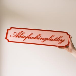 Puede incluir: Un letrero rectangular con un borde rojo y la palabra "Absofuckinglutely" en escritura cursiva roja. El letrero tiene un fondo rosa claro y está sostenido por una mano.