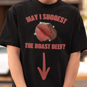 May include: A black short-sleeve t-shirt featuring the red text "MAY I SUGGEST THE ROAST BEEF?" above a graphic of a piece of roast beef with sparkles and a red downward-pointing arrow printed below the meat graphic on the front of the shirt.