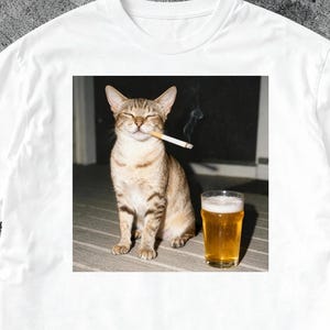 以下が含まれることがあります： タバコをくわえた猫とビールのグラスが描かれた、ユーモラスなイメージの白いTシャツ。猫は薄茶色で濃い縞模様。ビールは金色です。