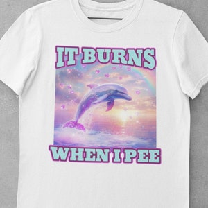 May include: White t-shirt with a graphic of a dolphin jumping out of the water with a rainbow and hearts. The text "IT BURNS WHEN I PEE" is printed below the graphic.