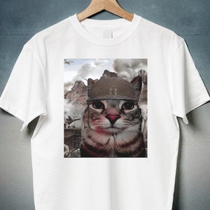 Puede incluir: Camiseta blanca con un gráfico de un gato con un casco militar. El gato está mirando directamente al espectador. El fondo es una imagen borrosa de una escena de guerra.