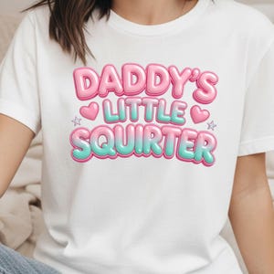 Puede incluir: Camiseta blanca con el texto "Daddy's Little Squirter" en letras de burbujas rosas y turquesas. El diseño incluye corazones y estrellas rosas. La persona lleva vaqueros azules.