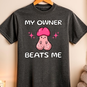 May include: Dark gray t-shirt with the text "MY OWNER BEATS ME" above a cartoon illustration of a penis with a pink head and pink star accents. The shirt is hanging on a wooden hanger.