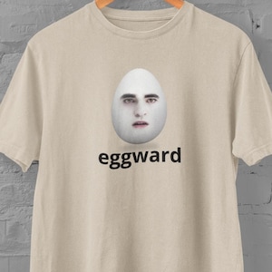 May include: A beige t-shirt with a white egg-shaped image of a person's face and the word "eggward" printed in black below the image.