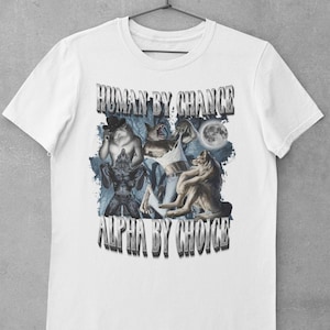 May include: A white short-sleeve t-shirt featuring a graphic print of multiple wolves and a full moon. The text "HUMAN BY CHANCE" appears at the top and "ALPHA BY CHOICE" at the bottom in a metallic, block-letter font. The design includes blue forest imagery.